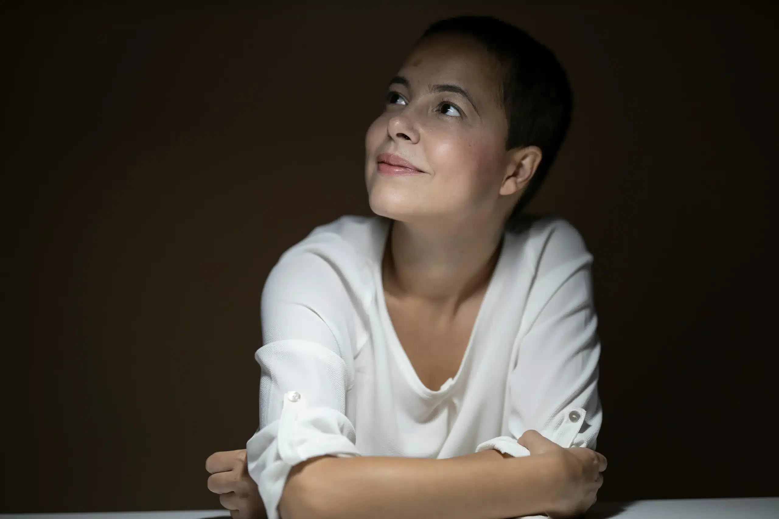 A woman with short hair wearing a white blouse sits at a table, looking up with a serene expression. The background is dark, creating a contrast that highlights her face in Andala Health Ketamine in San Antonio, TX.