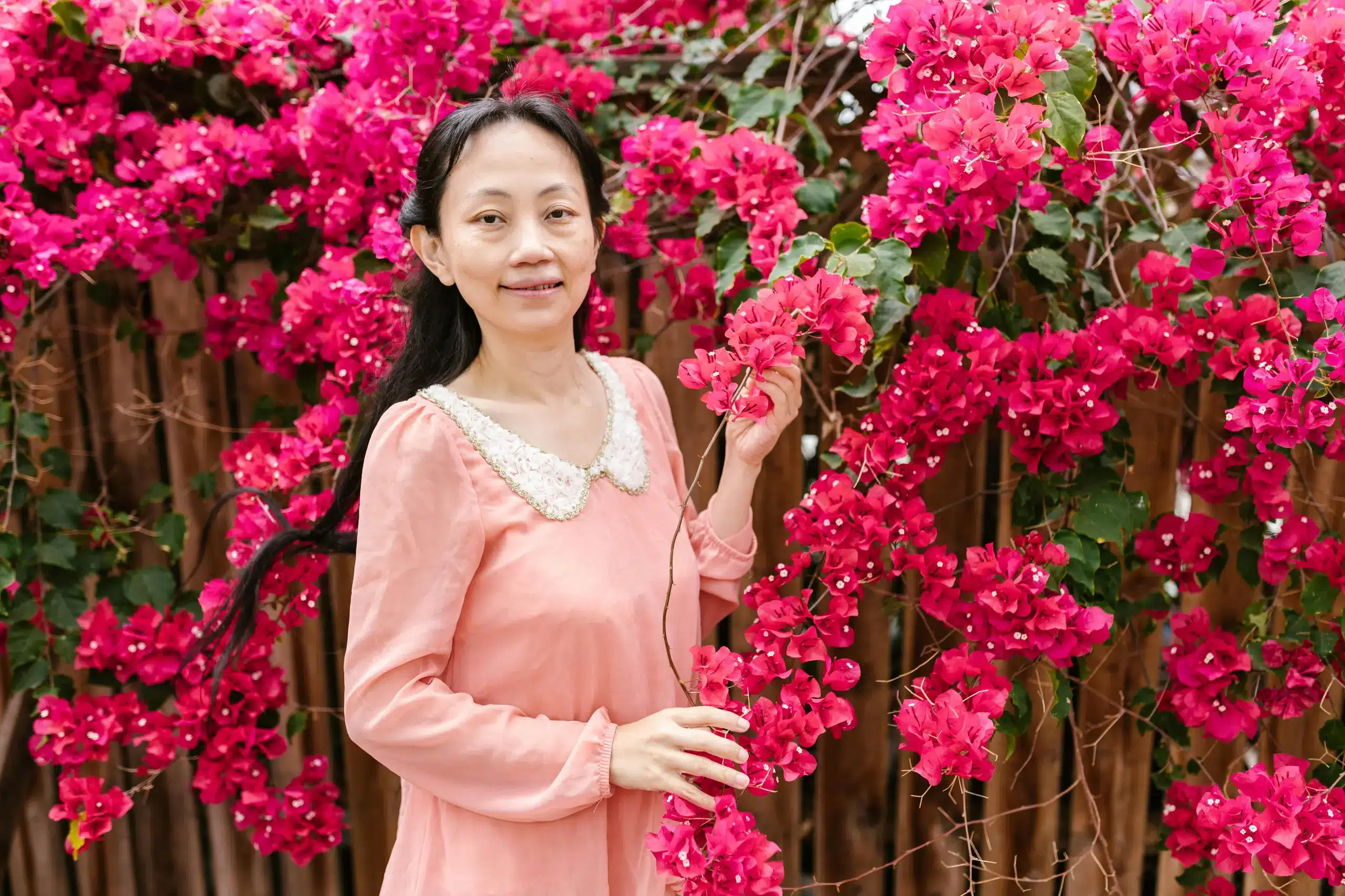A woman in a peach-colored dress with a lace collar stands amidst vibrant pink bougainvillea flowers, gently holding a branch while smiling softly in Andala Health Ketamine in San Antonio, TX.