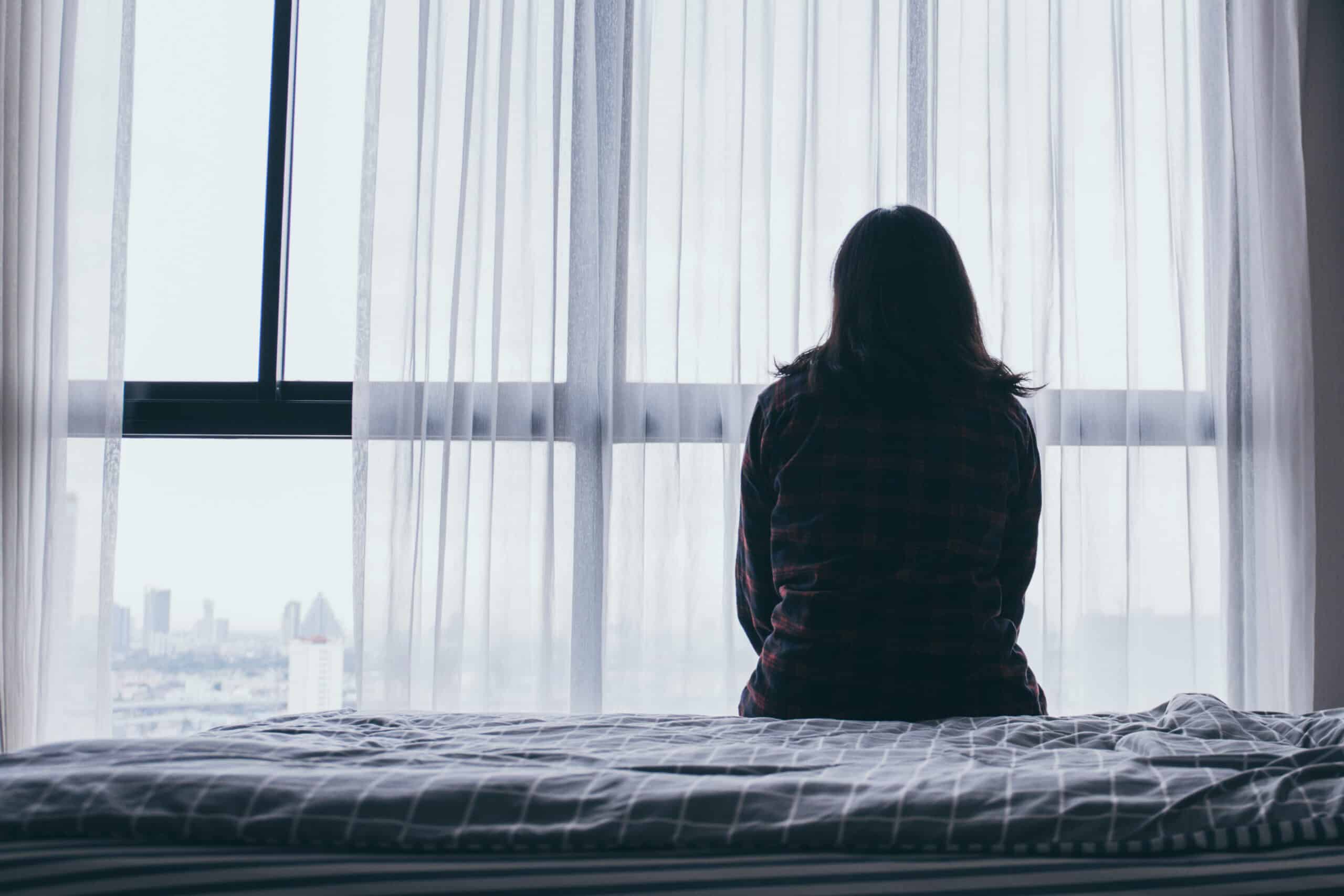 A woman sits on a bed looking out a window, representing feelings of depression and isolation. Used in a blog about Spravato treatment for major depressive disorder (MDD) and treatment-resistant depression (TRD).